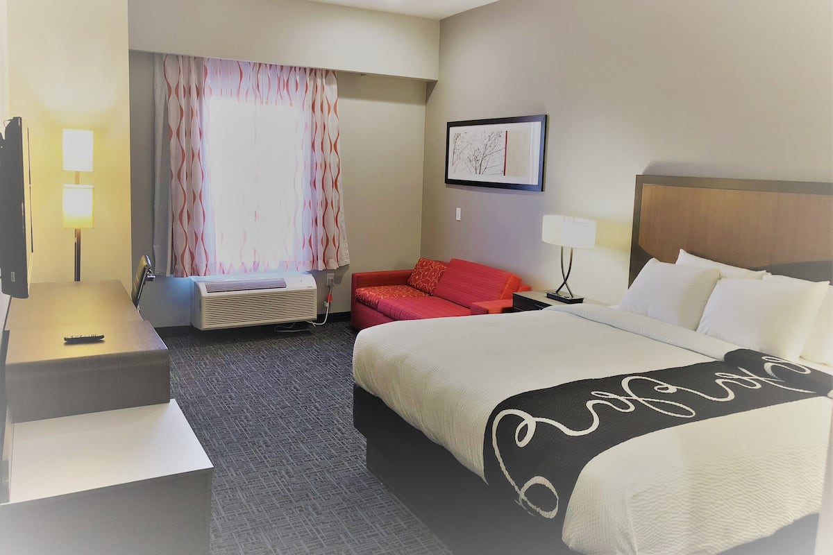 A hotel room with a bed, a desk, a chair, a TV, a lamp, a window with curtains, and a painting on the wall.