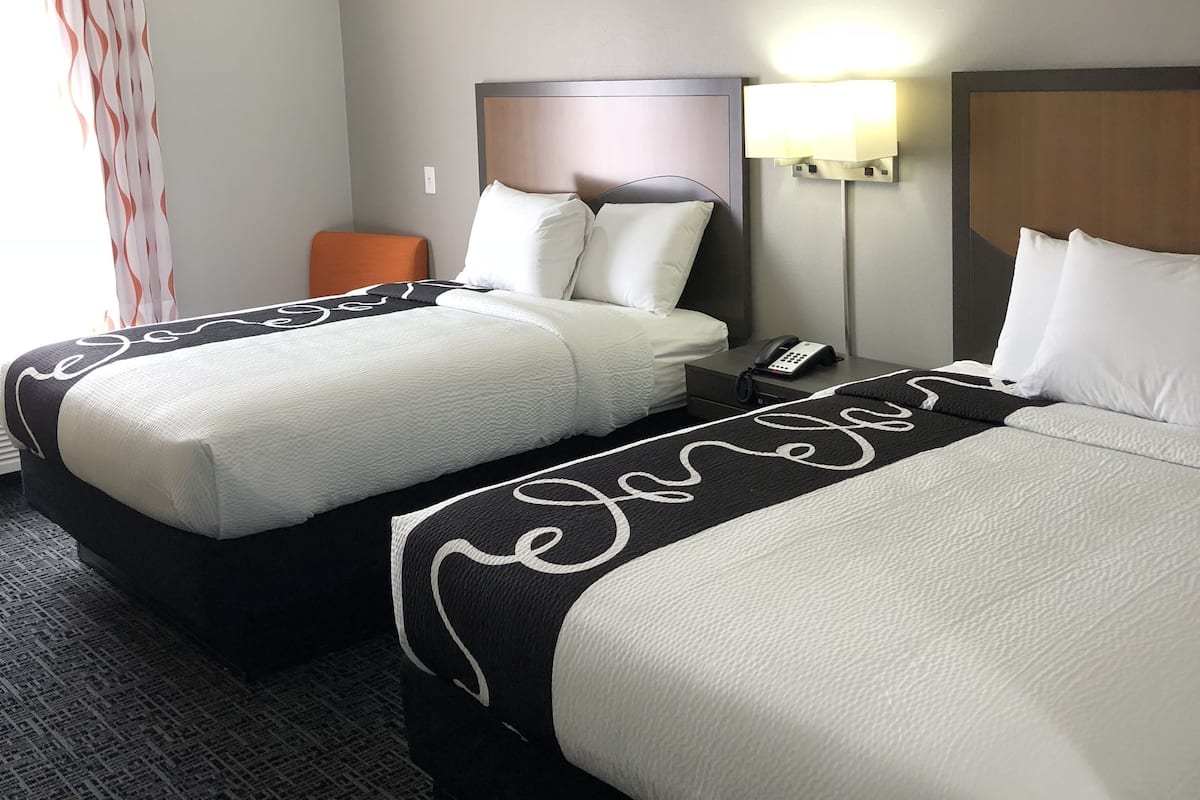 A hotel room with two beds, a nightstand, a lamp, and a chair.