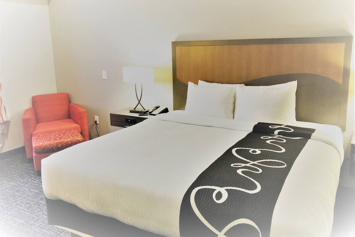 A hotel room with a large bed, a nightstand with a lamp, a red chair, and a wooden headboard.