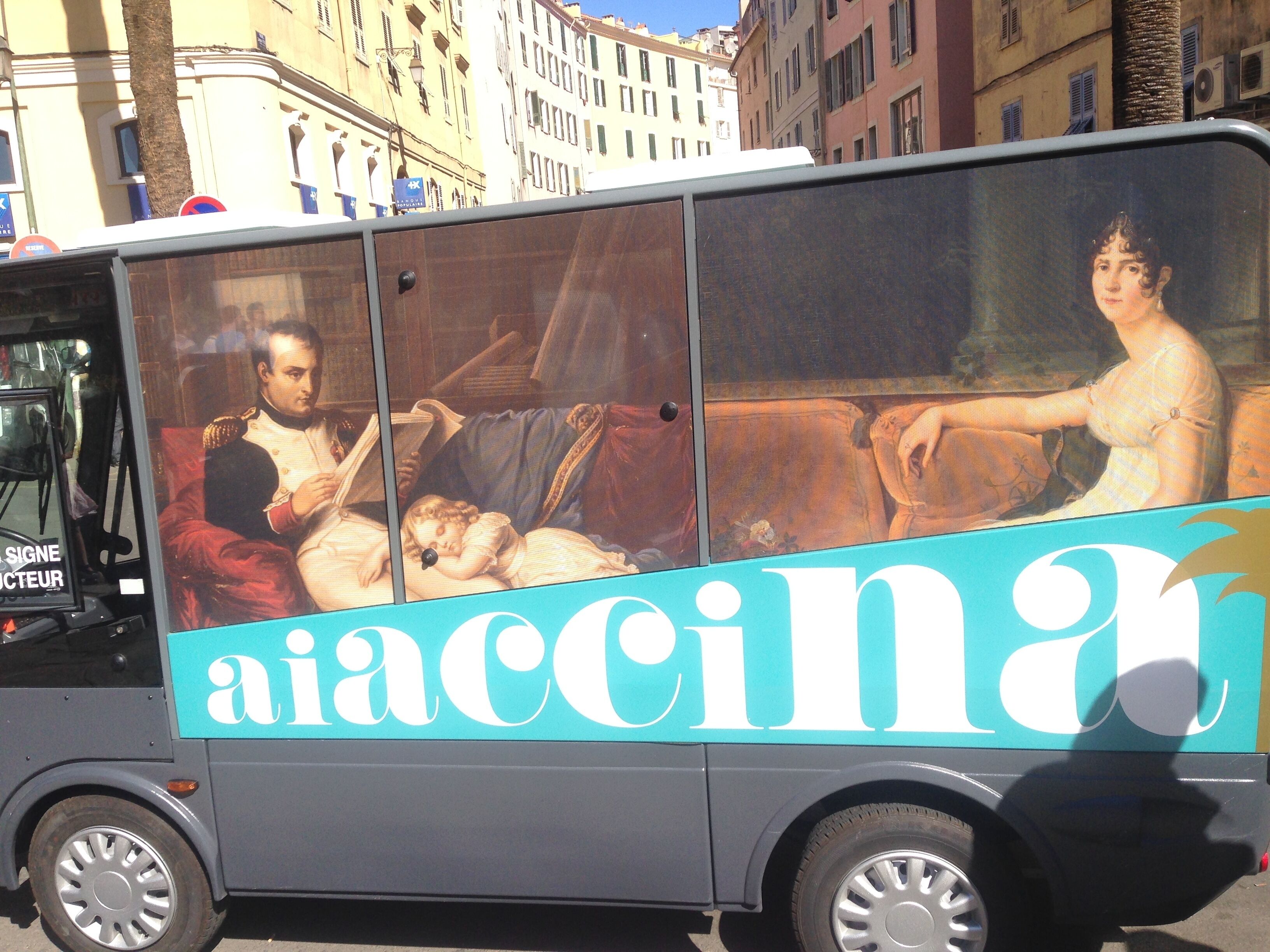 Ein Bus mit großflächiger Werbung, darunter ein Gemälde einer Mann und Frau, und dem Schriftzug 'aiaccina' an der Seite.