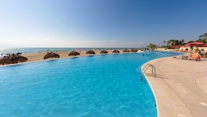 A swimming pool with a clear blue water, a sandy beach, and red umbrellas. - New Oceanfront Condo Grand Venetian (Puerto Vallarta)