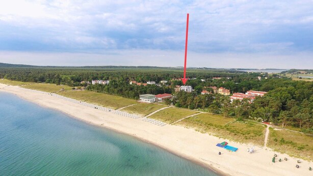 Vue aérienne d’une zone côtière avec une plage, une rangée de parasols et une rangée de bâtiments aux toits rouges.