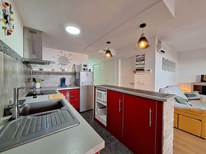 Une cuisine de taille réduite, équipée d’un évier, d’une cuisinière et d’un réfrigérateur. Les meubles sont de couleur rouge et il y a un petit coin repas avec une table et des chaises.