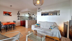 Ein modernes Wohnzimmer mit einer Couch, einem Glastisch, einer Essecke mit Tisch und Stühlen sowie einer Küche im Hintergrund.
