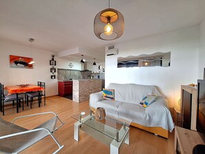 Un salon moderne avec un canapé, une table basse en verre, un coin repas avec une table et des chaises, et une cuisine en arrière-plan.
