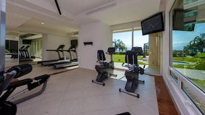 Ein Fitnessstudio mit Laufbändern, Crosstrainern und Gewichtbänken, große Fenster mit Blick auf Palmen und Stadtlandschaft.