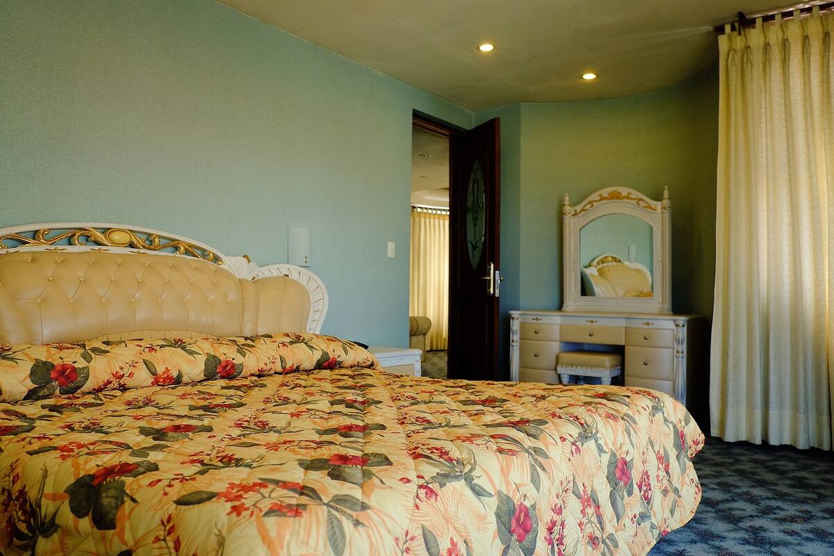 Ein Schlafzimmer mit Blumenoptik auf der Bettdecke, einem gepolsterten Kopfteil, einer Kommode mit Spiegel und einer Tür, die in einen weiteren Raum führt.