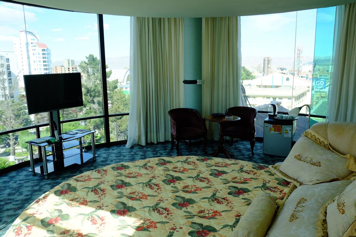 Ein Hotelzimmer mit einem großen Bett, einem Schreibtisch mit Computer, einem Sessel und einem Fernseher. Das Zimmer verfügt über große Fenster mit Blick auf die Stadt.