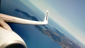 Ala de avión con el texto "RYANAIR" y vista de una costa que muestra una combinación de paisajes urbanos y naturales.