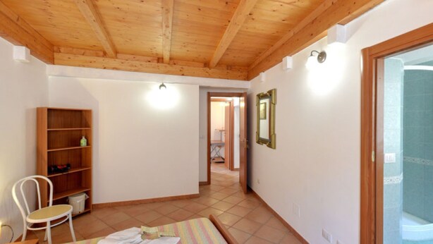 A room with a wooden ceiling, a white chair, a shelf with items, a bed with a striped blanket, and a door leading to a bathroom with a shower. - Casa Machin Alghero (Alghero)