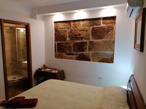 A bedroom with a bed, a stone wall, a small table with items, and a bathroom visible through a glass door. - Casa Machin Alghero (Alghero)