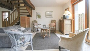 Een gezellige woonkamer met een grijze bank, rieten stoelen, een kleine ronde tafel en een trap die naar een hoger niveau leidt.