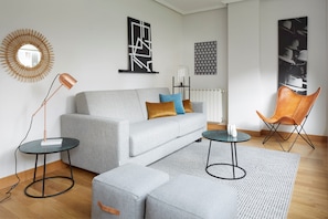 A modern living room with a grey sofa, two side tables, a coffee table, a patterned rug, a wooden floor, and a wall-mounted mirror.