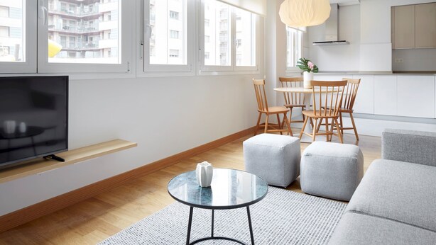 Ett modernt vardagsrum med en TV, ett runt bord i glas, en grå soffa, trästolar och en matplats med ett vitt bord och stolar.