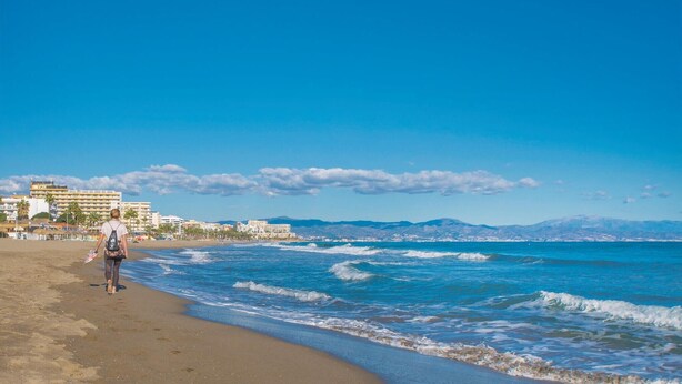 Una playa con una persona caminando por la orilla, edificios al fondo y un cielo azul despejado.