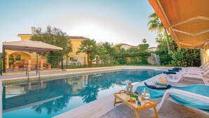 Piscina con sillones de descanso y una mesa con bebidas, rodeada de vegetación y edificios.