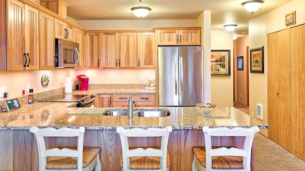 A kitchen with wooden cabinets, stainless steel appliances, a granite countertop, and a seating area with white chairs. - Alpine Villagers (Leavenworth)