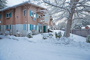 A two-story wooden house with a green door and windows, surrounded by snow-covered ground and trees.