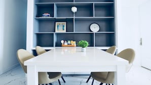 Un comedor moderno con una mesa blanca, sillas color beige, una lámpara colgante azul y un estante gris con objetos decorativos.