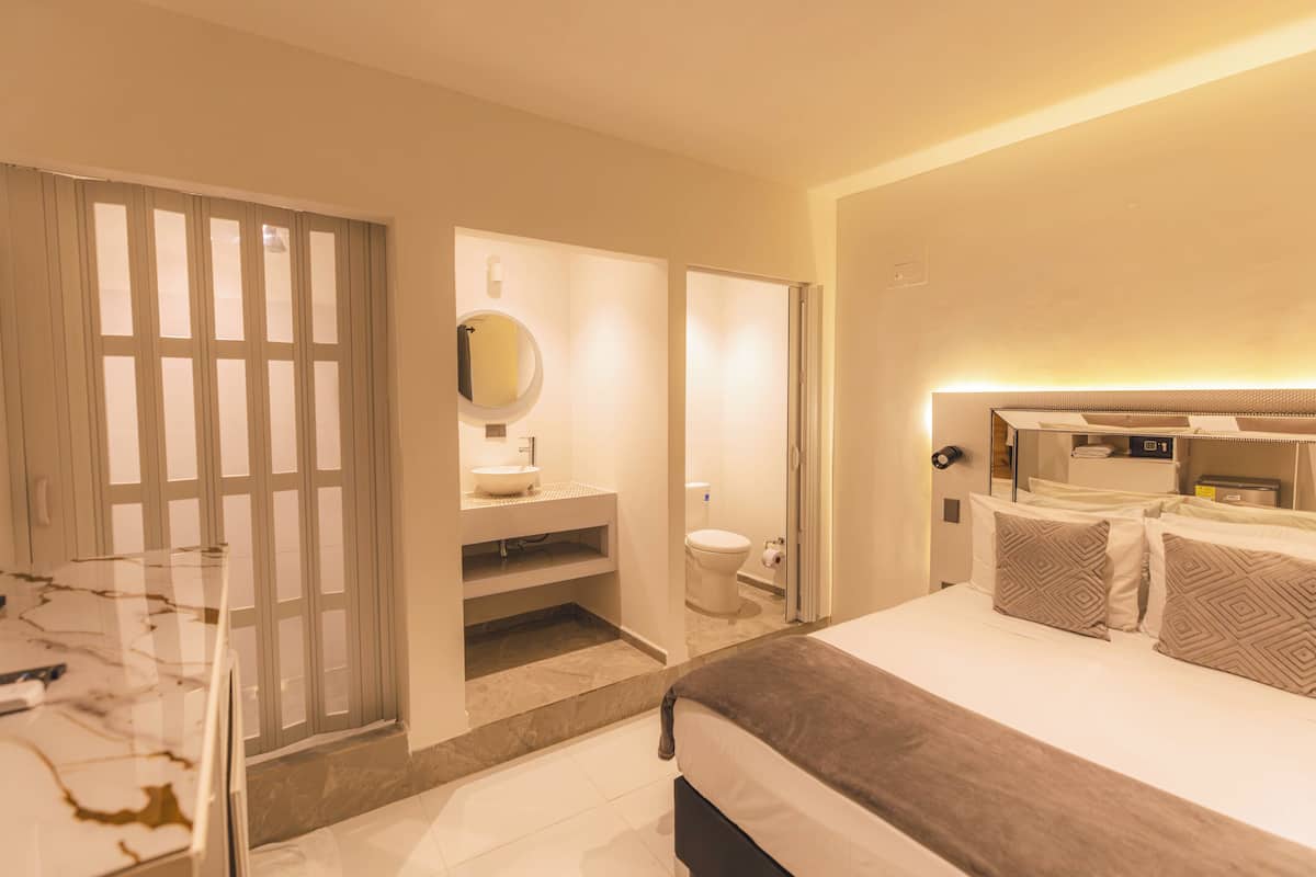 A modern hotel room with a large bed, a bathroom with a toilet and sink, and a glass partition separating the bedroom from the bathroom.