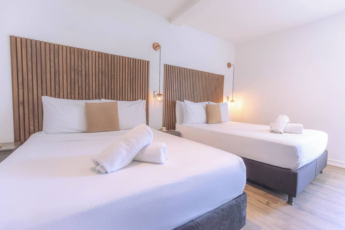 Two beds with white linens and wooden headboards, a small table with a cup, and a wall-mounted light fixture.