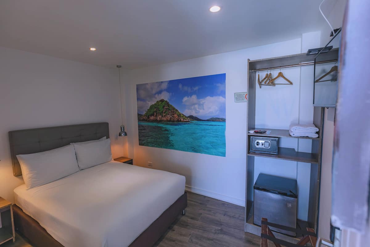 A bedroom with a bed, a nightstand, a wall-mounted radio, a shelf with towels, and a painting of a coastal scene.