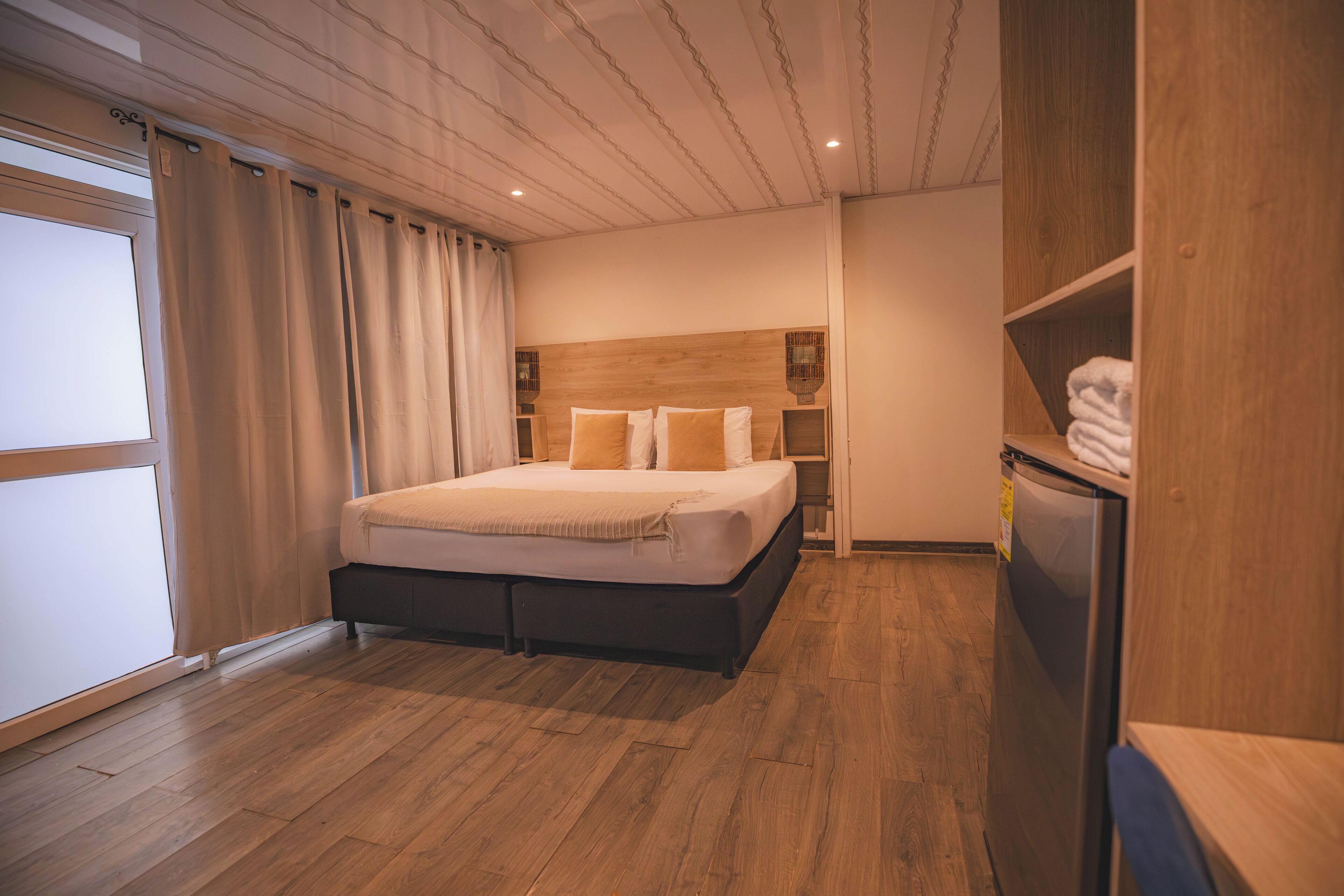 Quarto com piso de madeira, cama com roupa de cama branca e bege, cabeceira de madeira e guarda-roupa embutido com espelho.
