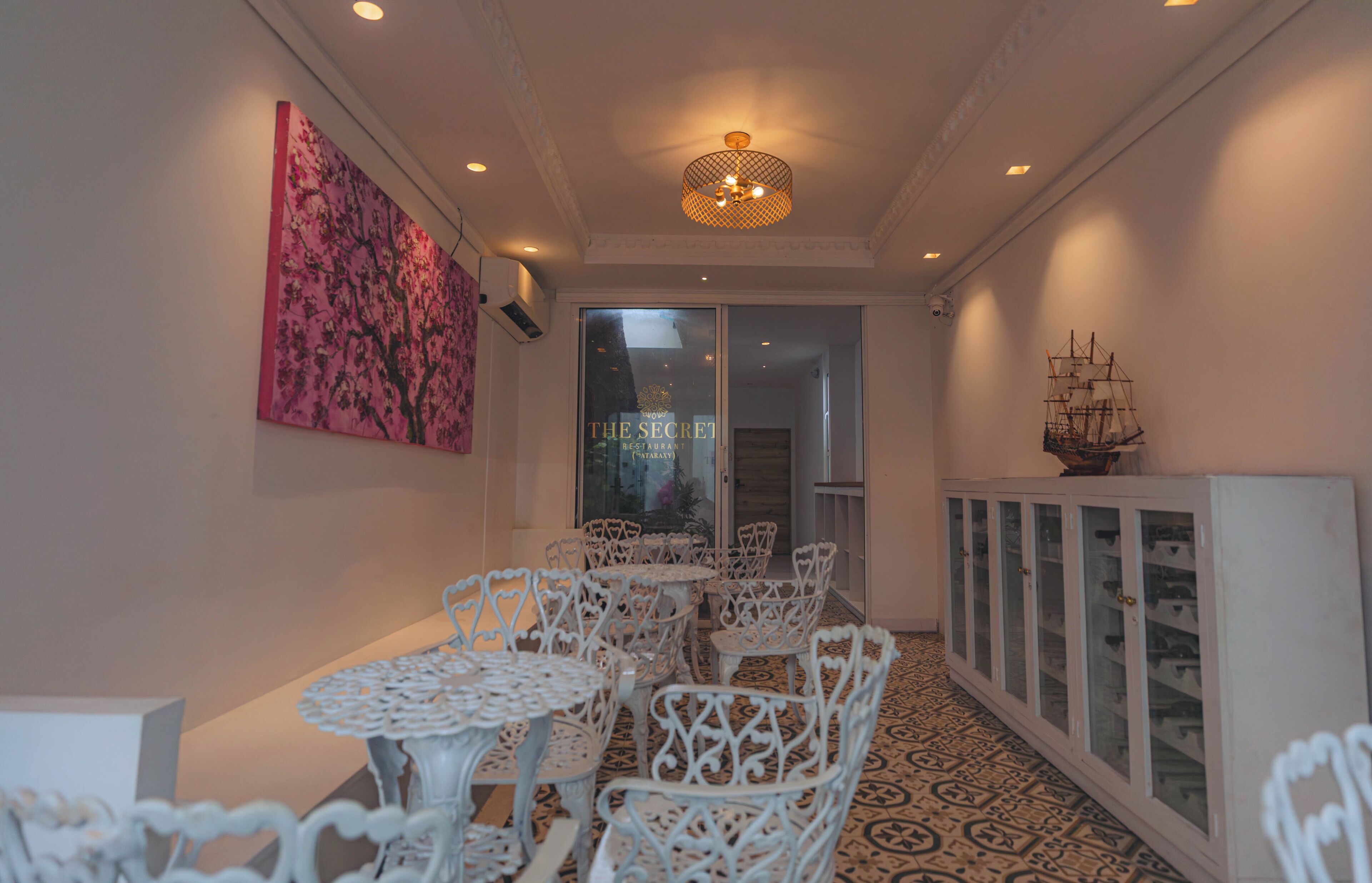 A corridor with white ornate chairs and tables, a patterned floor, a ship model on a cabinet, and a pink artwork on the wall.