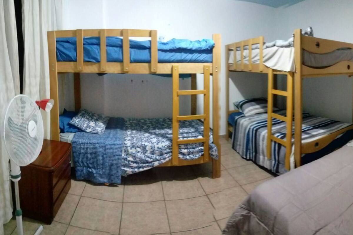 Une chambre avec deux lits superposés, une commode en bois, un ventilateur et un rideau.