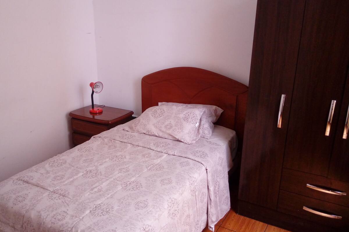 Un petit lit bien fait, avec une couvre-lit à motifs, une table de chevet surmontée d’une lampe rouge et un porte-manteau adossé à un mur uni.