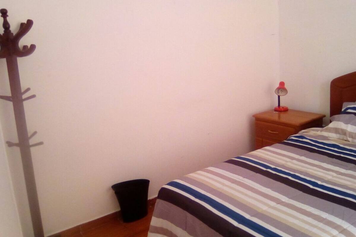 Une chambre à coucher avec un lit, une table de chevet, une lampe, un porte-manteau et une poubelle.