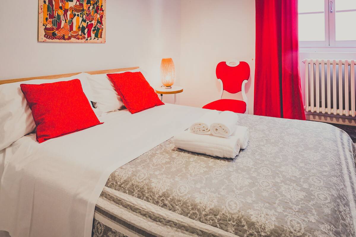 A neatly made bed with red and white bedding, a small table with a lamp, and a red chair in a room with a radiator and a window.