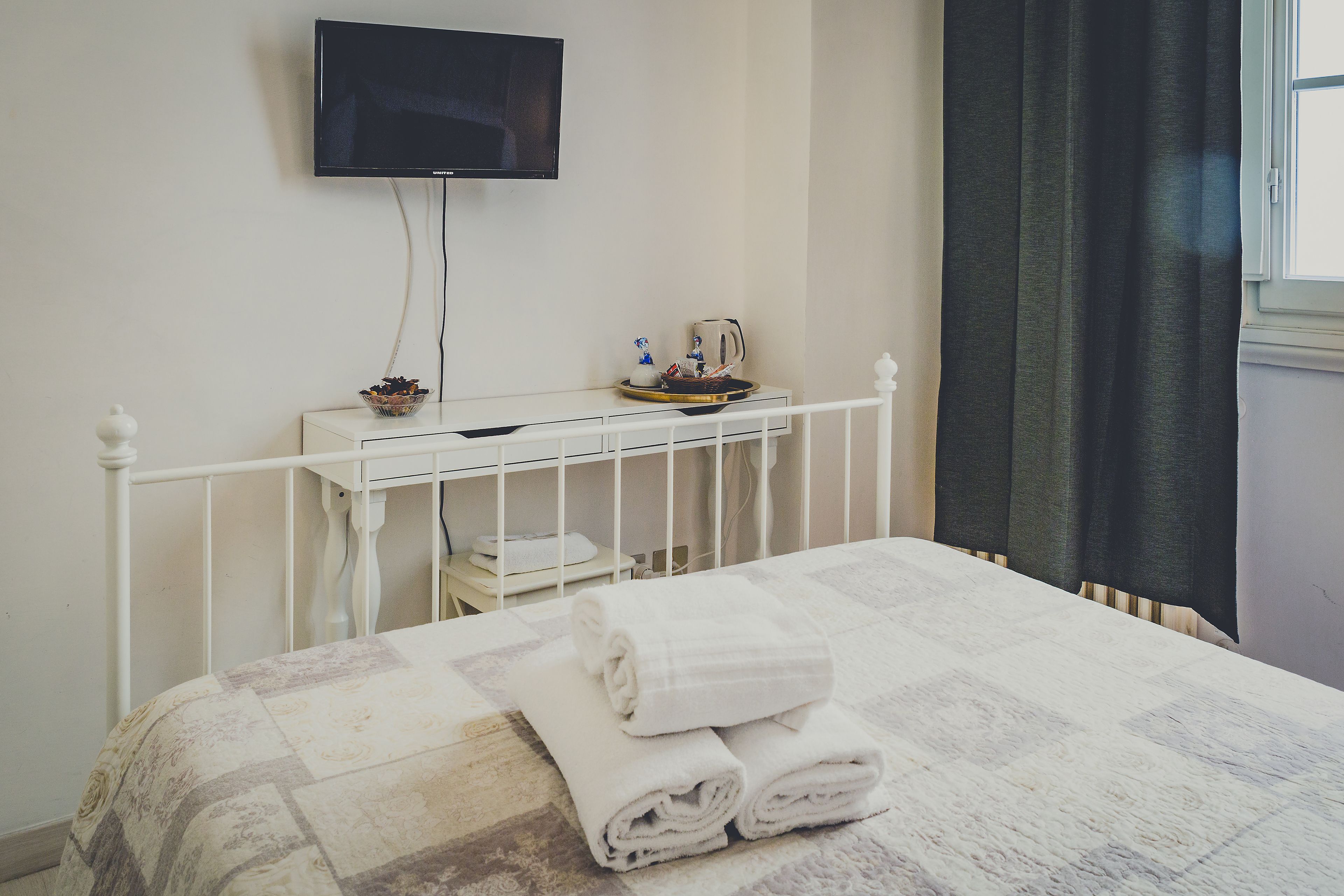 A neatly made bed with folded towels, a bedside table with a bowl of fruit, a kettle, and a cup. A television is mounted on the wall above the table.