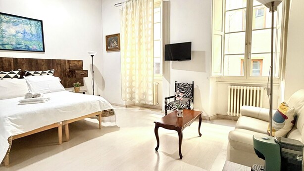 A spacious room with a bed, a chair, a table, and a sofa. There is a window with curtains, a painting on the wall, and a radiator. - Il Gattopardo (Florence)