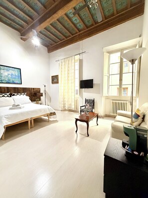 A spacious room with a bed, a chair, a table, and a sofa. There is a window with curtains, a painting on the wall, and a radiator. - Il Gattopardo (Florence)