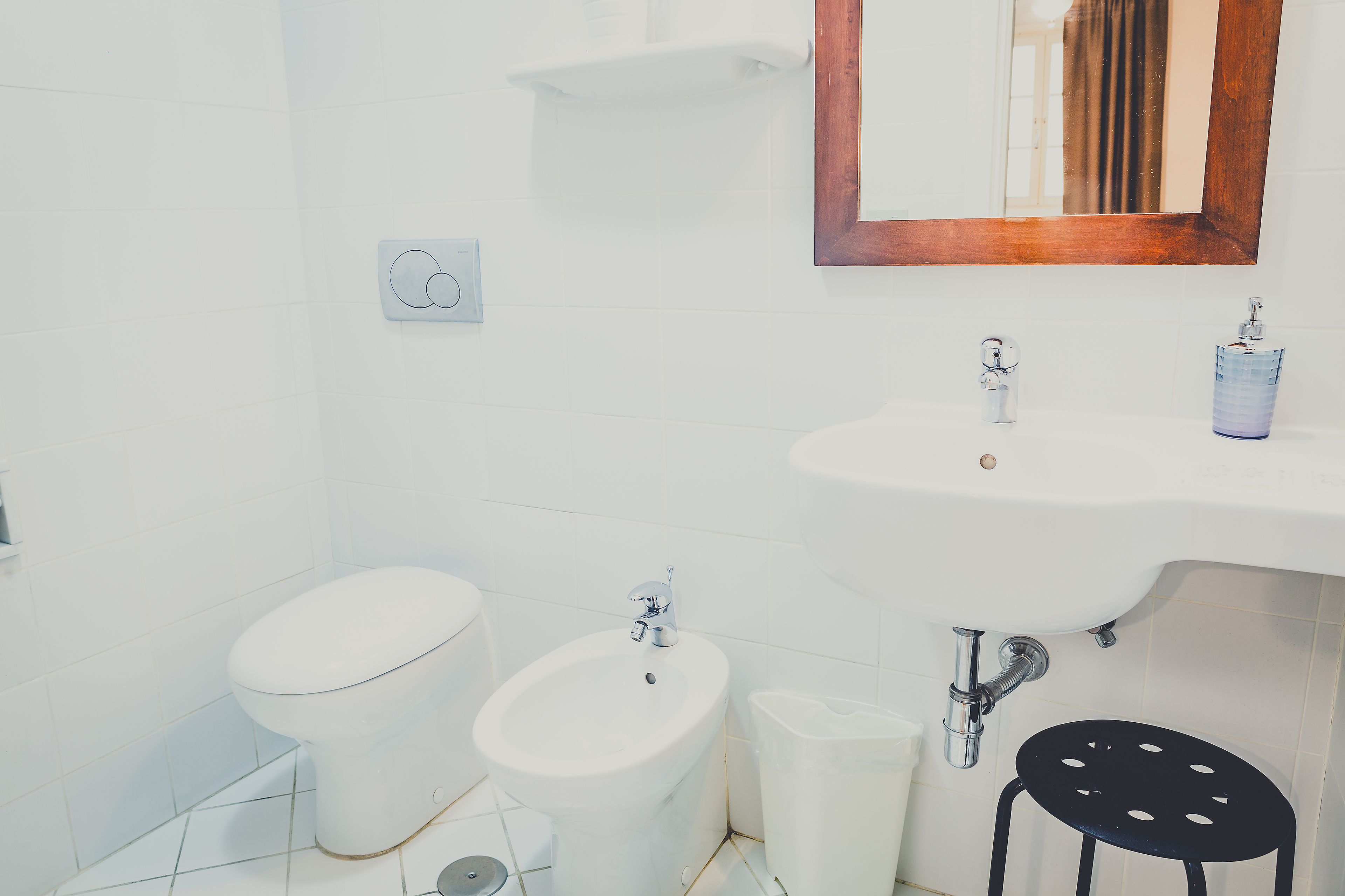 A bathroom with a toilet, bidet, sink, mirror, and a black stool.
