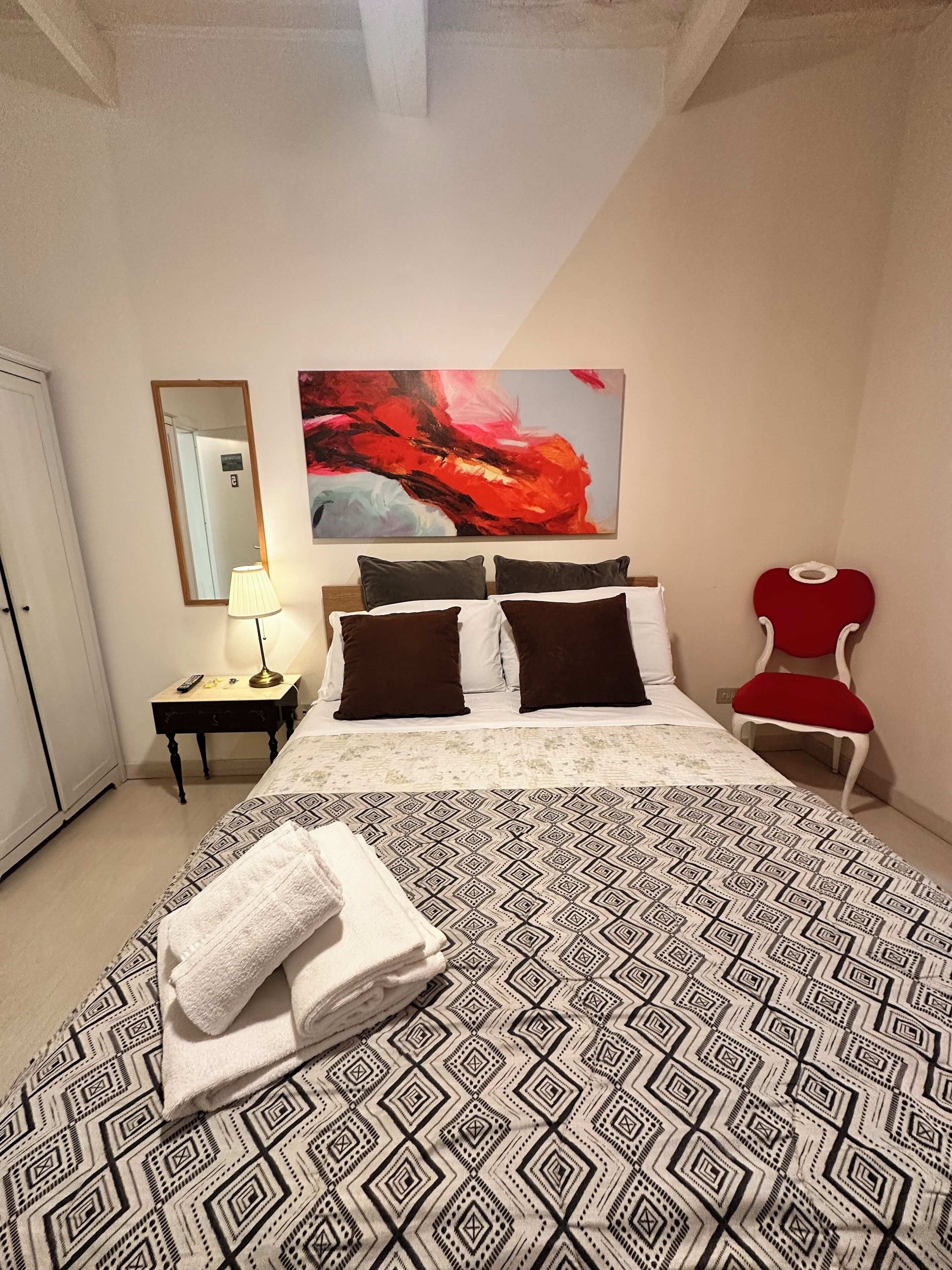 A neatly arranged bedroom with a large bed, a bedside table with a lamp, a red chair, and a large abstract painting on the wall.