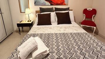 A neatly arranged bedroom with a large bed, a bedside table with a lamp, a red chair, and a large abstract painting on the wall.