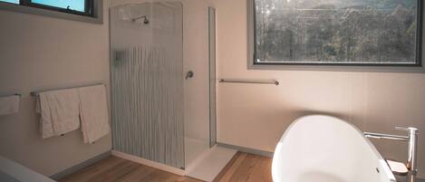 A modern bathroom with a glass shower enclosure, a freestanding bathtub, and a window offering a view of a forested hillside.