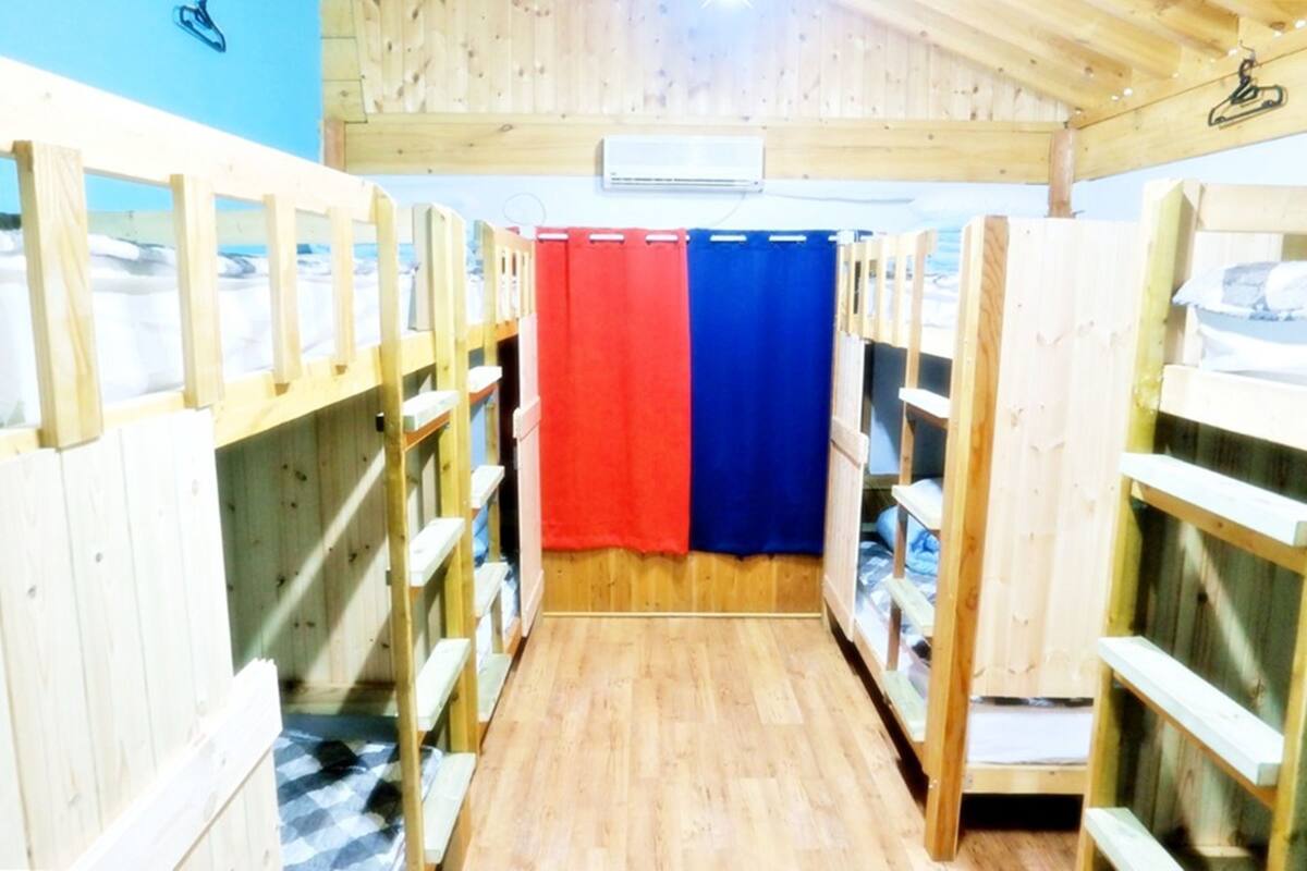 A row of wooden bunk beds with a checkered floor, a red and blue curtain, and a ceiling fan.