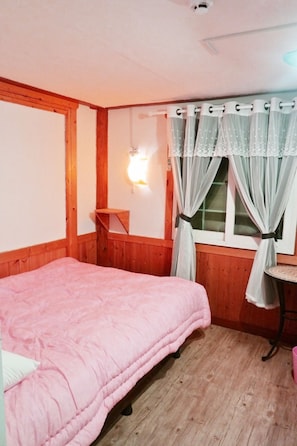 Een slaapkamer met een houten bed, een nachtkastje, een raam met gordijnen en een wandlamp.