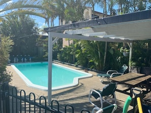 Una piscina con borde blanco, rodeada de palmeras y un área de estar cubierta con mesas y sillas.