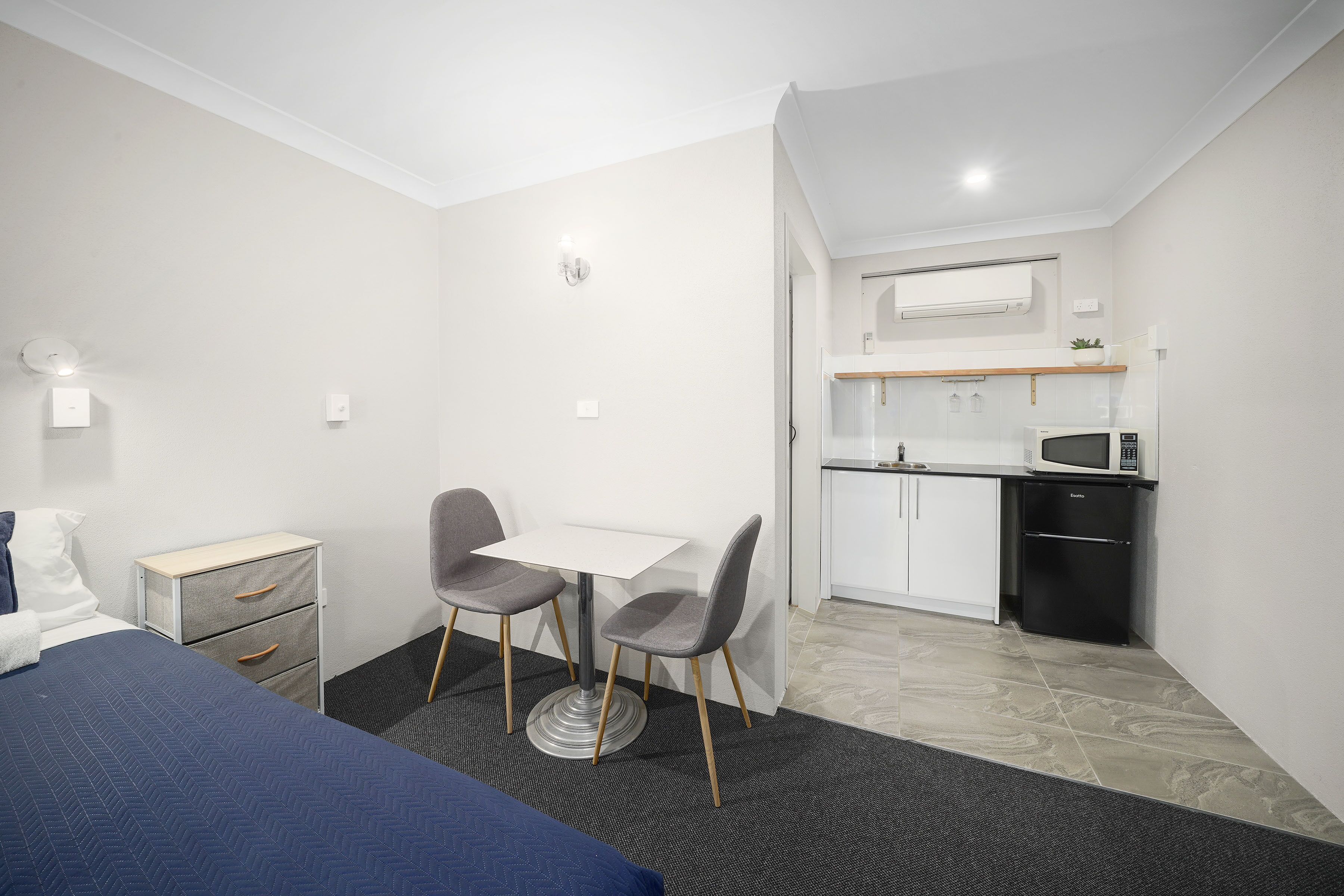 A compact hotel room with a bed, a desk with two chairs, a small kitchenette, and a bathroom.