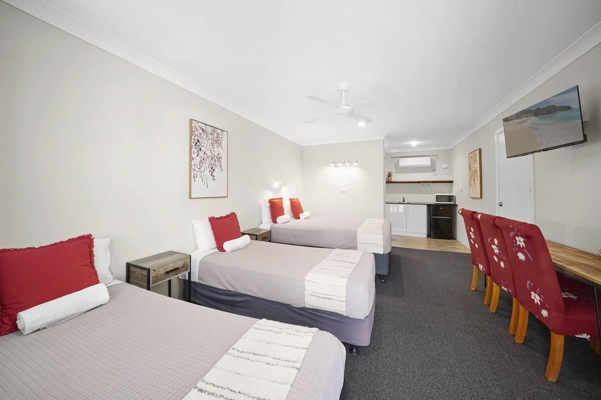 A hotel room with two beds, a desk, chairs, a TV, and a kitchenette.