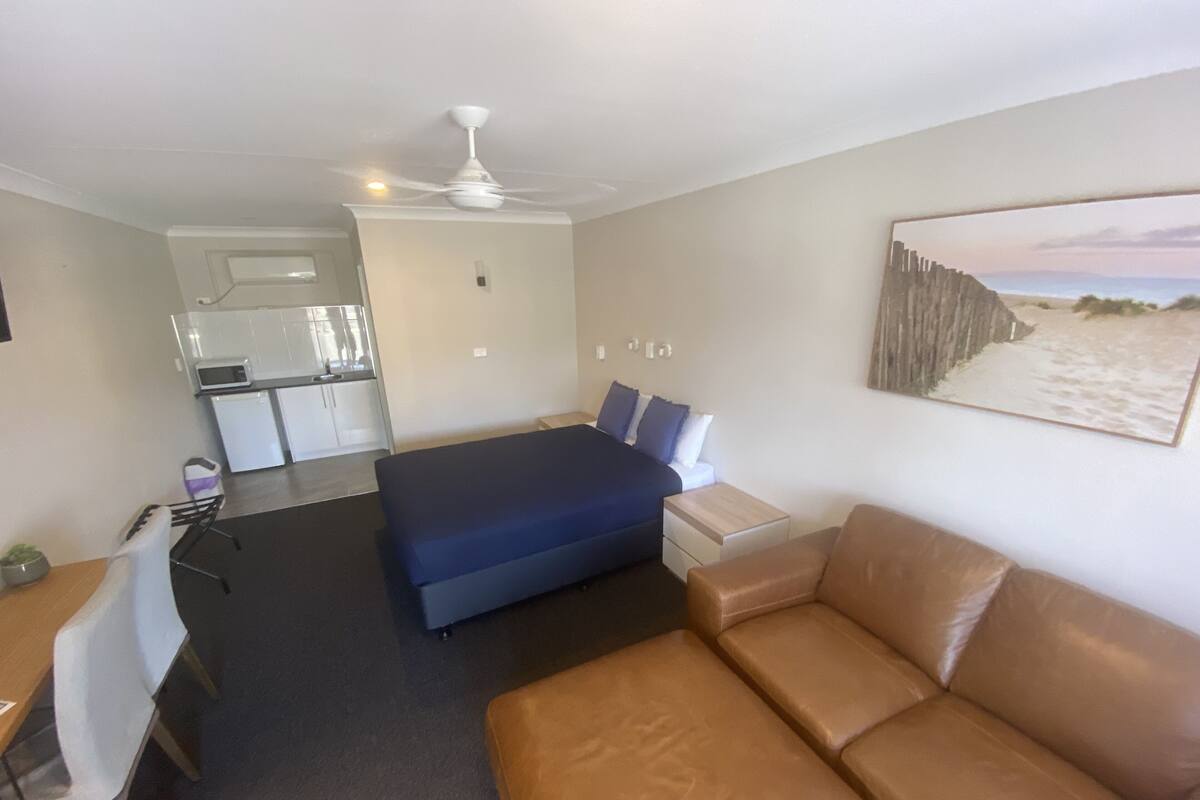 A bedroom with a bed, a brown leather couch, a desk, a chair, a kitchenette, and a ceiling fan.