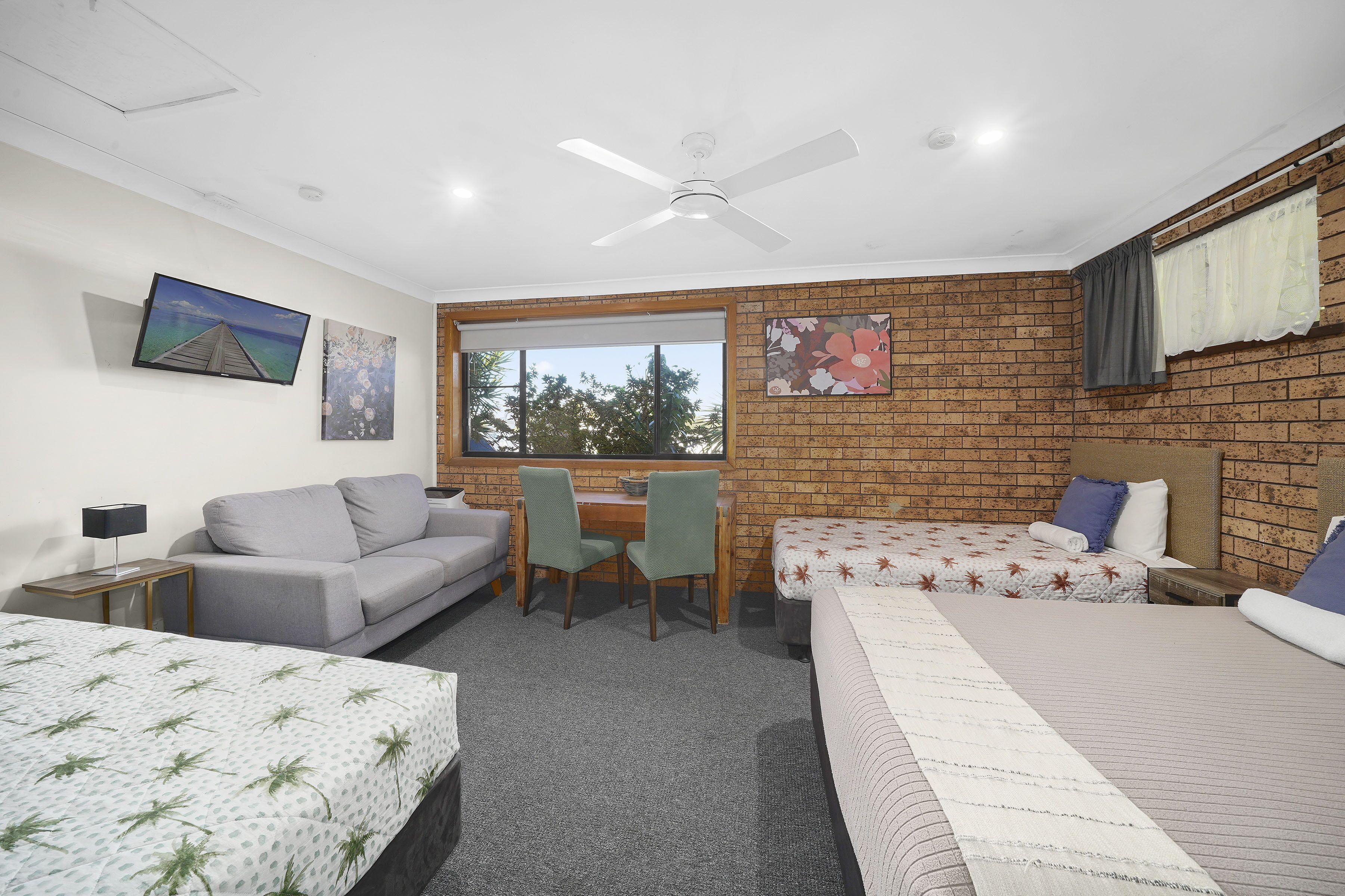 A room with a brick wall, a desk, two beds, a sofa, and a ceiling fan.