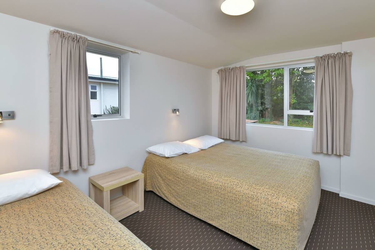 Quarto com duas camas, uma mesa de cabeceira de madeira pequena, uma janela com cortinas e uma luminária no teto.