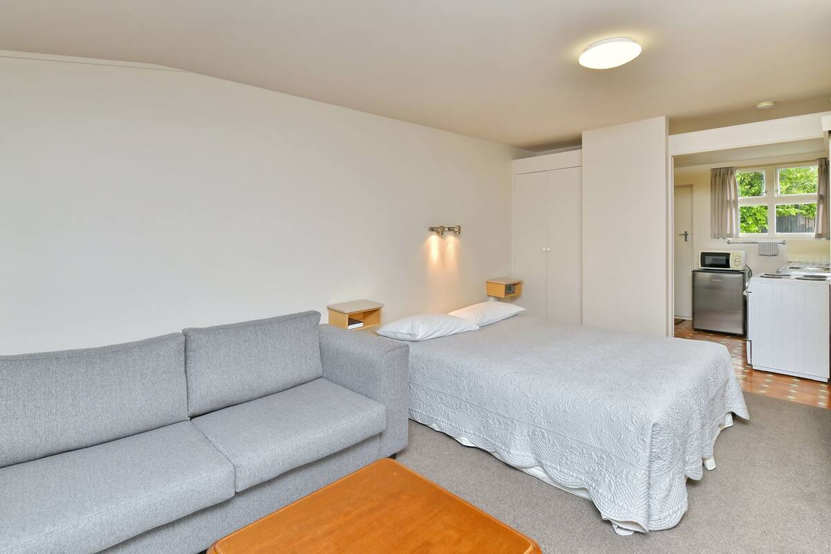 Quarto com um sofá cinzento, uma cama com colcha branca, uma pequena mesa de madeira e uma kitchenette ao fundo.