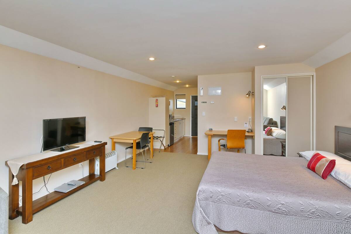 Quarto de hotel com uma cama, escrivaninha, cadeira, televisão e cozinha compacta.