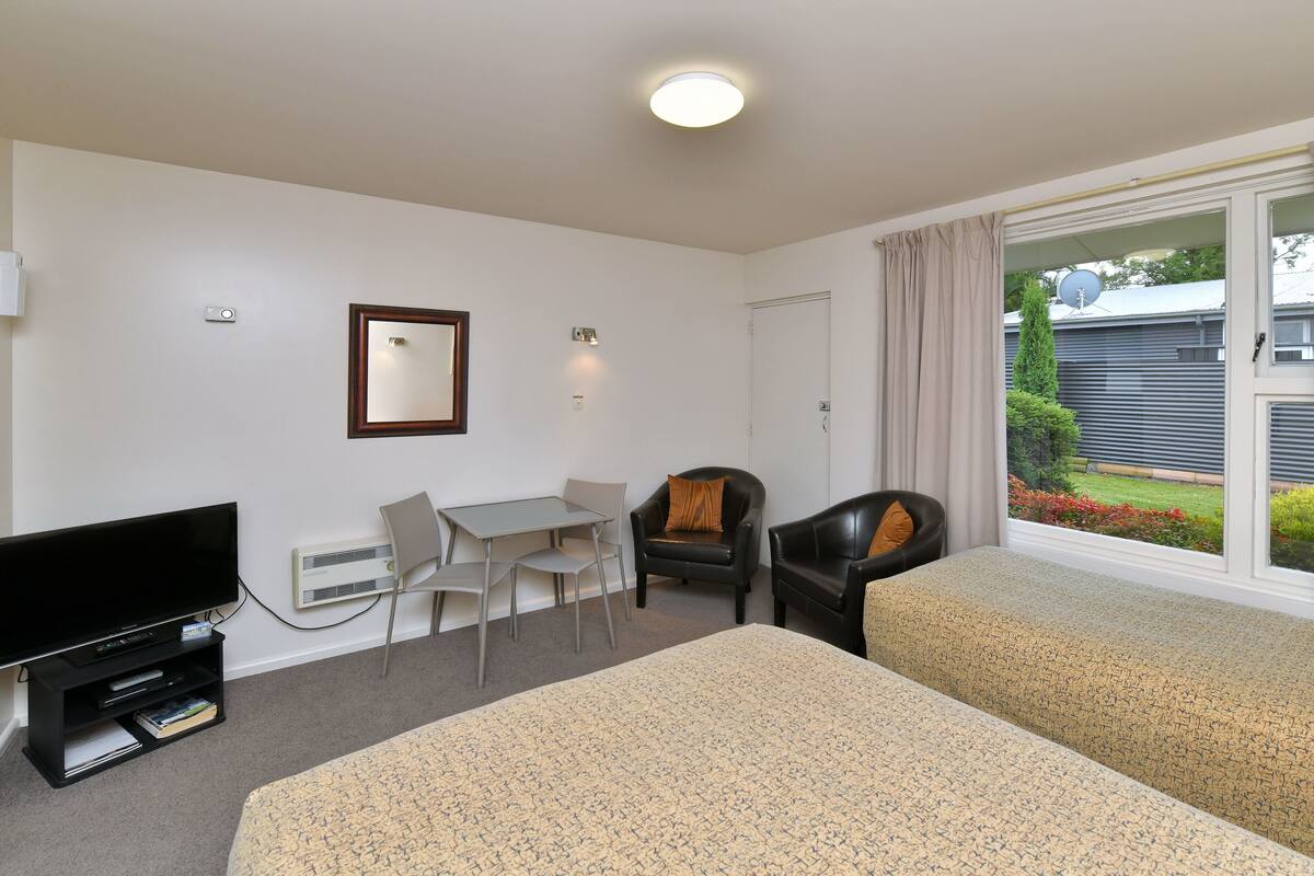 Quarto de hotel com cama, escrivaninha, cadeira, televisão e janela com vista para uma casa e jardim.